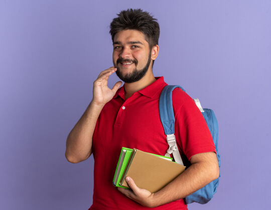 人年轻的留着胡子的学生 穿着红色马球衫 背着书包 脸上带着羞涩的微笑 站在蓝色的墙上害羞年轻马球