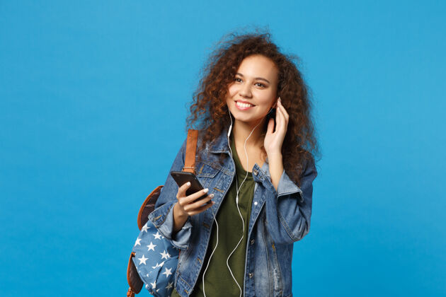 耳机年轻的非洲裔美国少女 穿着牛仔服 背着耳机 隔离在蓝色的墙上人女人享受