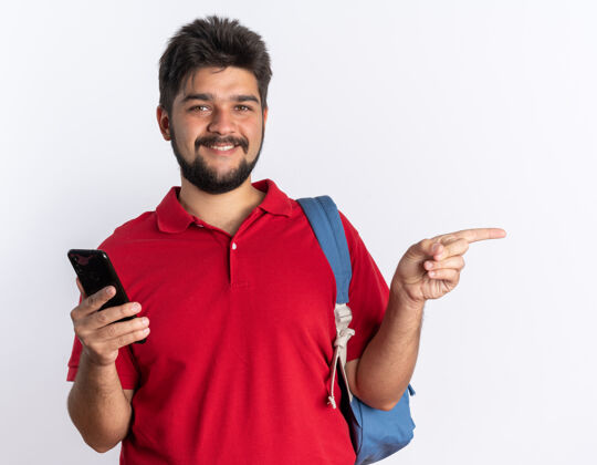 指向年轻的留着胡子的学生 穿着红色马球衫 背着背包 拿着智能手机 食指指向一边 微笑着自信地站在白墙上姿势站马球