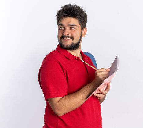 手势年轻的留着胡子的学生 穿着红色马球衫 背着背包 站在白色的墙上 在笔记本上写着自信的微笑站立笔记本姿势