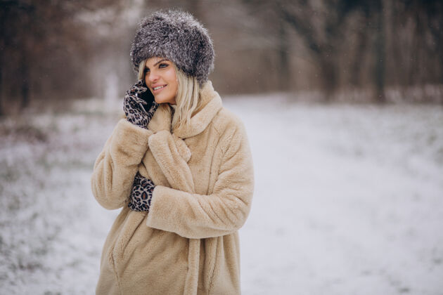 寒冷穿着冬衣的女人走在满是雪的公园里打电话毛衣美丽帽子