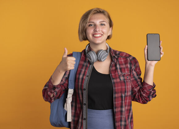 向上微笑的年轻斯拉夫女学生戴着耳机 背着背包 竖起手机大拇指拇指女孩背包