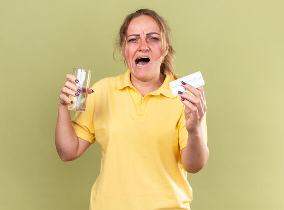 抱着一个穿着黄色衬衫的不健康的女人站在绿色的墙上 手里拿着一杯水和感冒药 感觉很糟糕站着感冒不健康