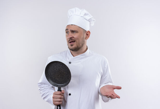 壁板迷茫的年轻帅哥厨师身着厨师制服 手里拿着煎锅 两手空空地看着隔离在白墙上的一面手空困惑