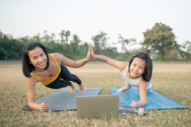积极妈妈和女儿在做瑜伽在公园里训练妇女和儿童户外运动健康的运动生活方式 chaturanga姿势健康 正念概念 在笔记本电脑上看在线视频教程孩子草地健康