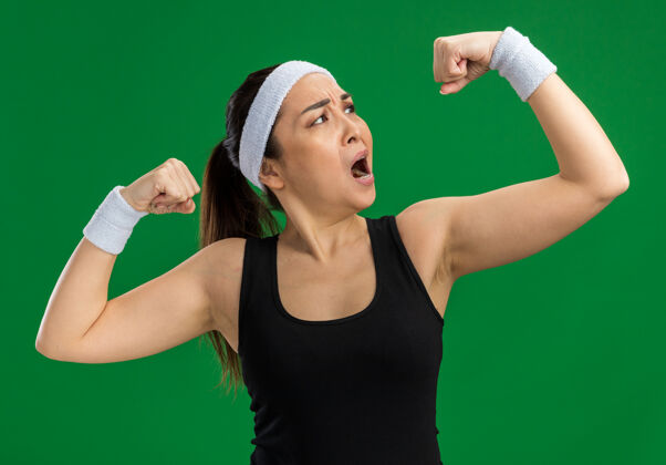 女子戴着头带和臂章的年轻健身女士看起来绷紧了举起的拳头 二头肌站在绿色的墙上站立拳年轻