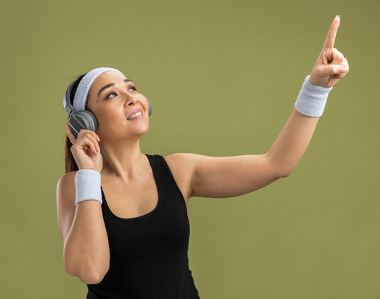 站立戴着头带和耳机的年轻健身女士站在绿色的墙上 用食指指着她最喜欢的音乐微笑着指点最爱音乐