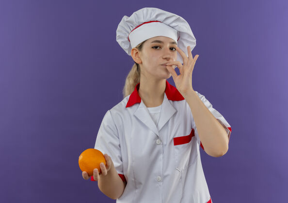 年轻自信的年轻漂亮厨师穿着厨师制服做着可口的手势 拿着橘子看起来孤立在紫色的墙上美味手持制服