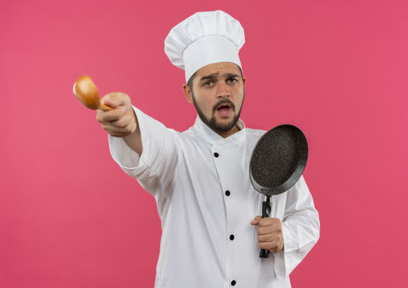 烹饪穿着厨师制服的年轻男厨师手拿煎锅 伸出汤匙 孤立地站在粉红色的墙上薯条制服厨师
