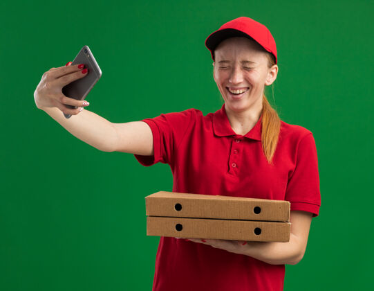 拿着身穿红色制服 头戴鸭舌帽 拿着比萨饼盒 用智能手机自拍的年轻送货女孩站在绿色的墙壁上开心地微笑着帽子欢呼盒子