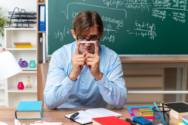 年轻年轻的男老师戴着眼镜 拿着粉笔 神情严肃地坐在课桌旁 教室的黑板前放着书和笔记戴着教室男