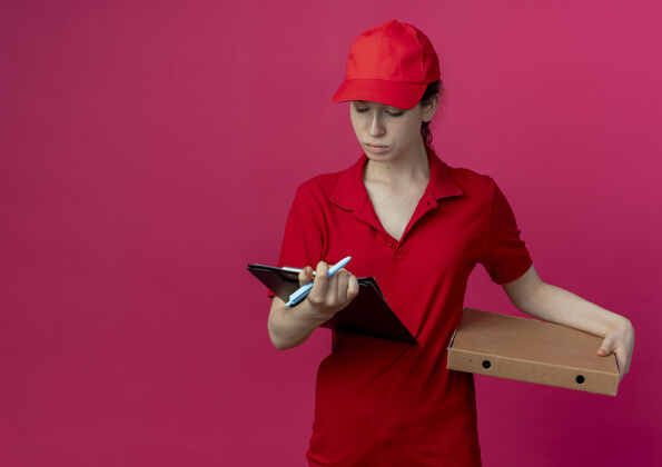 空间年轻漂亮的送外卖的女孩 穿着红色制服 戴着帽子 手里拿着比萨饼包装笔和剪贴板 看着剪贴板送货持有包装