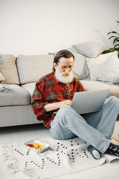 舒适长相不错的老人用笔记本电脑工作坐在地板上吃快餐观看下载个人