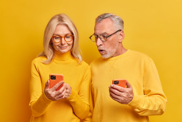 阅读震惊的胡须老人盯着现代智能手机设备 显示妻子使用现代小玩意在互联网上阅读令人震惊的新闻隔离黄墙在线妻子年龄