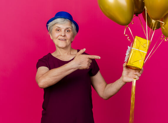 点戴着派对帽的快乐的老妇人手持氦气球 指着粉红色的礼盒礼物穿帽子