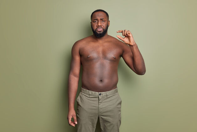 衣服不高兴的留着胡子的美国黑人男子裸体摆姿势展示小东西姿势展示非常小的物体对着深绿色的墙摆姿势躯干手身体