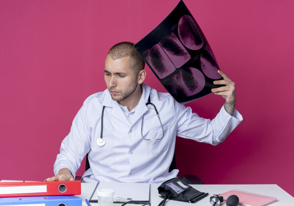 男性体贴的年轻男性医生 穿着医用长袍 手持听诊器 坐在办公桌旁 拿着工作工具 拿着x光片 看着隔离在粉红色墙上的办公桌长袍衣服年轻人