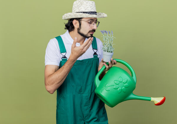 人年轻的留着胡须的园丁 穿着连体衣 戴着帽子 手里拿着浇水罐和盆栽植物 站在淡绿色的墙上 感受着宜人的气味壶浇水衣服