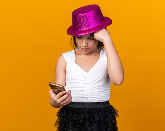 电话焦急的年轻白人女孩 戴着紫色的派对帽 把拳头放在额头上 拿着手机 看着隔离在橙色墙上的手机 还有复印空间年轻白种头