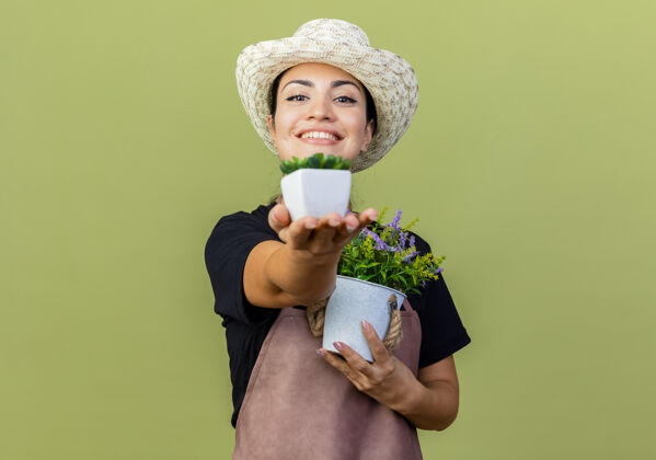 光年轻漂亮的女园丁 围着围裙 戴着帽子 手里拿着盆栽植物 站在淡绿色的墙边 兴高采烈地微笑着看着前方围裙花园人