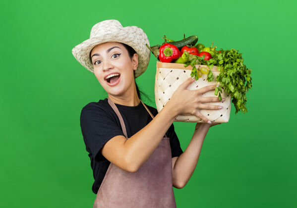市民年轻漂亮的女园丁围着围裙 戴着帽子 手里拿着装满蔬菜的箱子 站在绿色的墙上 面带微笑地看着前面帽子围裙箱子