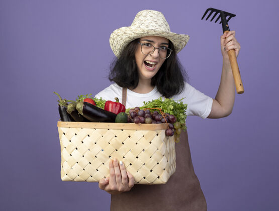 人愤怒的年轻黑发女园丁戴着眼镜 穿着制服 戴着园艺帽 把菜篮子和耙子孤立在紫色的墙上耙子篮子帽子