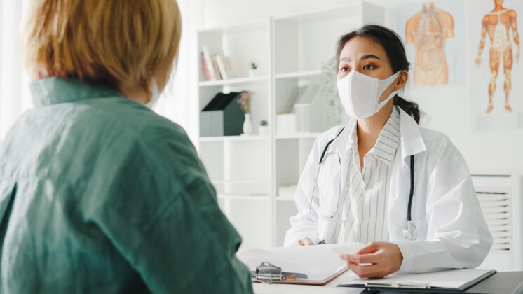 预防年轻的亚洲女医生戴着防护面罩 用剪贴板和医院办公室的女病人讨论结果或症状房间距离面罩