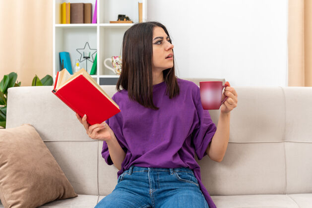 沙发穿着休闲服的年轻女孩拿着一本书和一杯茶 困惑地坐在客厅里的沙发上房间住靠边站