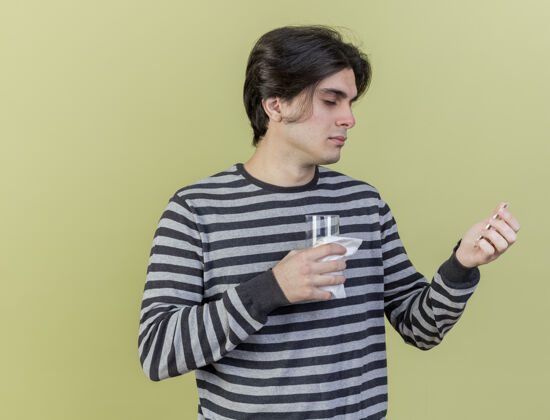 玻璃一个拿着药片的年轻人 用一杯水隔离在橄榄绿的pensativo上持有思考水