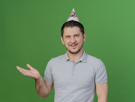 壁板微笑的年轻人戴着生日帽点着生日礼物 手放在绿色隔离的一边帽子年轻生日
