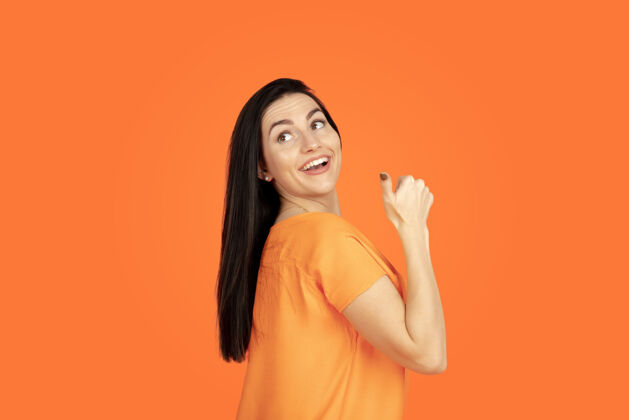 人橙色工作室背景上的白种人年轻女子肖像穿着衬衫的漂亮黑发女性模特人类情感的概念 面部表情 销售 广告广告空间指向 展示 微笑身长财务公司