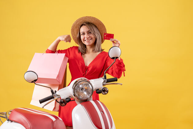购物正前方是手持购物袋和购物卡 穿着红色连衣裙 坐在轻便摩托车上的快乐女人轻便摩托车包运动