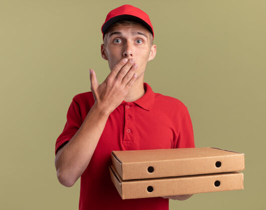 嘴巴焦急的年轻金发送货员把手放在嘴里 拿着橄榄绿的比萨饼盒焦虑男孩手