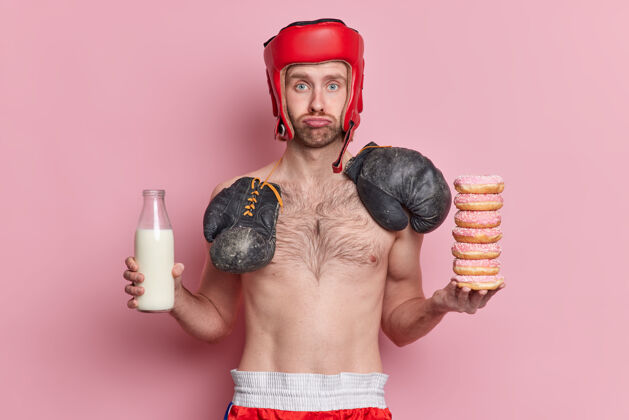 堆一个瘦骨嶙峋的男拳击手戴着帽子 脖子上的拳击林里放着一堆甜甜圈和一瓶牛奶手套不快乐粉色