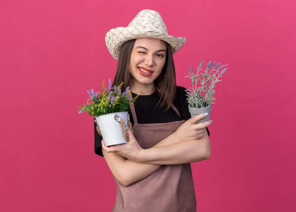 花园自信漂亮的白人女园丁戴着园艺帽眨着眼睛 双手交叉站在粉红色的花盆上站立自信帽子
