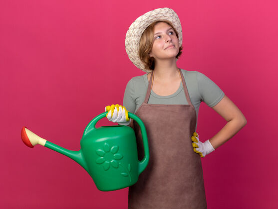 请年轻的斯拉夫女园丁高兴地戴着园艺帽和手套 手里拿着浇水罐 边看边上的粉红色壁板花园帽子