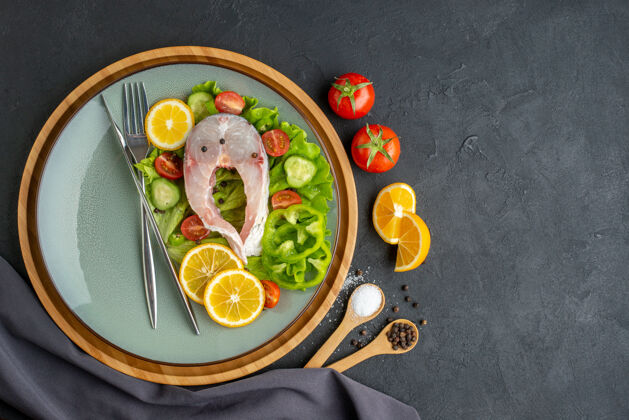 生鱼生鱼片和新鲜蔬菜的俯视图柠檬片和餐具放在灰色盘子上香料深色毛巾放在黑色表面的左侧壁板蔬菜顶部