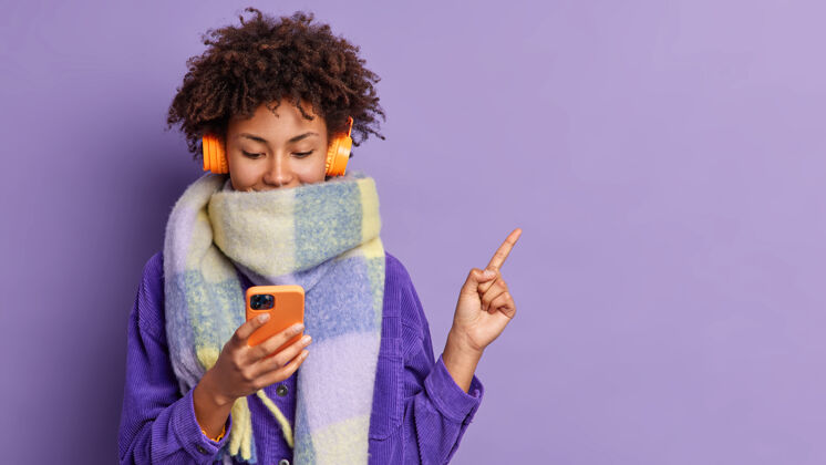 休闲年轻可爱的卷发少女拿着手机看游戏它选歌听右上角指示为寒冷的冬天戴围巾在紫色墙上显示复制空间立场女性电话