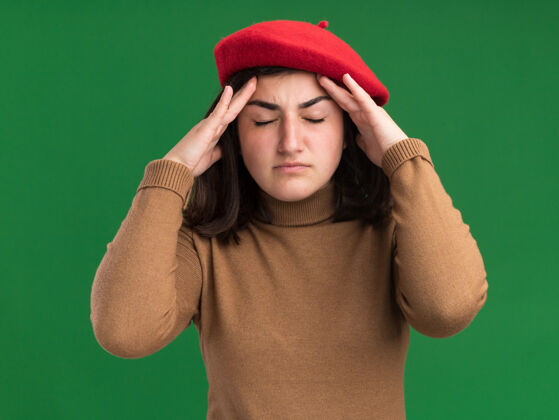 帽子戴着贝雷帽的年轻漂亮白人女孩把双手放在绿色的额头上前额绿色疼