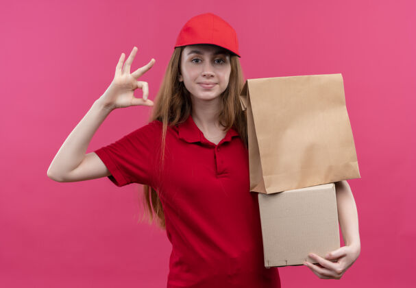 持有自信的年轻送货女孩穿着红色制服拿着箱子 在孤立的粉色空间上做着ok的标志信心制服好