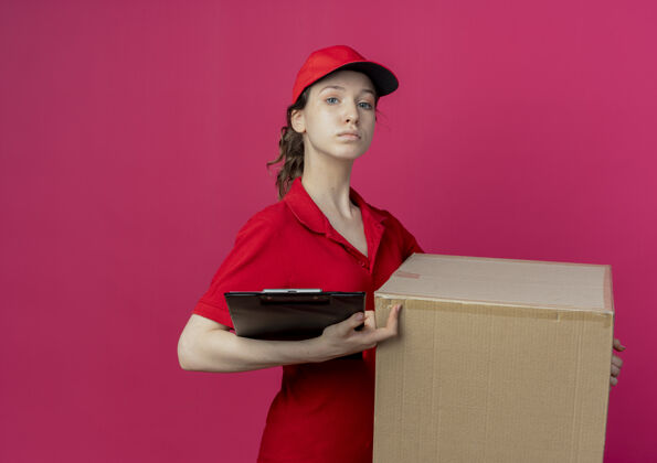 红色自信的年轻漂亮的送货女孩 穿着红色制服 戴着帽子 拿着纸盒和剪贴板 背景是深红色 有复印空间持有背景制服