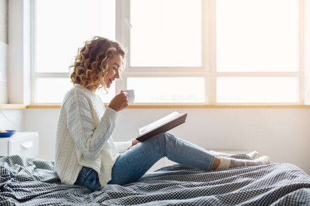 时尚美丽的年轻女子早上坐在床上看书 穿着白色针织毛衣 喝着咖啡房子房间女性