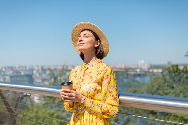 旅游穿着黄色夏装 戴着黄色帽子 喝着咖啡 享受阳光的女人的户外肖像 站在桥上 城市的景色令人惊叹年轻人城市景观欢乐