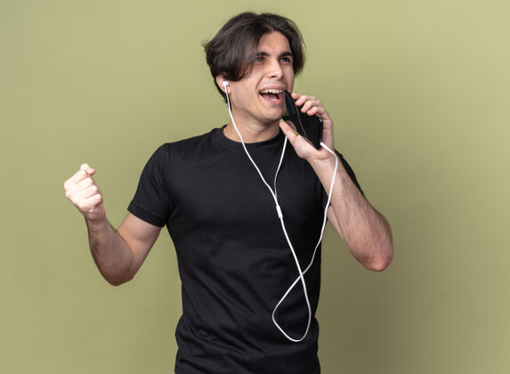 站着带着微笑的年轻帅哥 穿着黑色t恤 戴着耳机 拿着电话 在橄榄绿的墙上独自歌唱男人人感觉