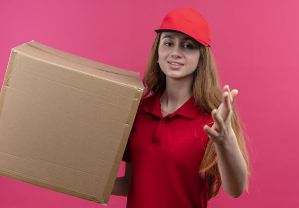 年轻自信的年轻送货女孩 穿着红色制服 拿着盒子 在孤立的粉色空间里伸出手来红色交货盒子