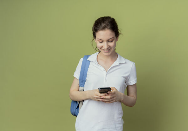 橄榄微笑的年轻漂亮的女学生戴着背袋用她的手机孤立在橄榄绿的背景与复制空间学生女性手机