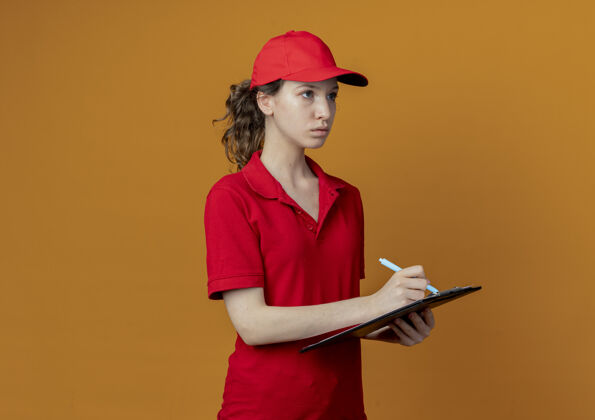 钢笔年轻漂亮的送货女孩 穿着红色制服 戴着帽子 拿着剪贴板和笔 笔直地看着 准备在橙色背景上孤立地写 还有复印空间帽子橙色送货