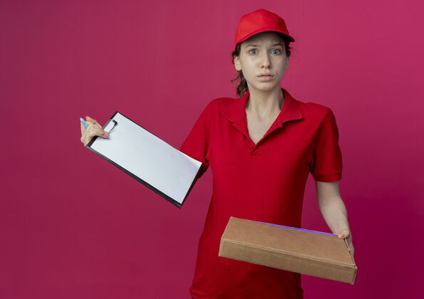 送货令人印象深刻的年轻漂亮的送货女孩在红色制服和帽子举行比萨饼包装笔和剪贴板孤立的深红色背景深红色披萨年轻