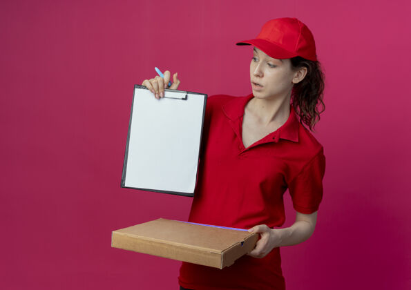 女孩令人印象深刻的年轻漂亮的送货女孩在红色制服和帽子拿着比萨饼包装笔和显示剪贴板看着剪贴板在深红色背景与复制空间隔离深红色红色印象