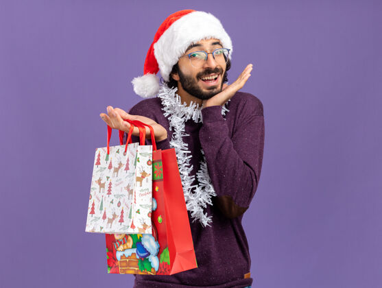 手兴奋的年轻帅哥戴着圣诞帽 脖子上戴着花环 手里拿着礼品袋 双手分开放在蓝色背景上包圣诞节礼物